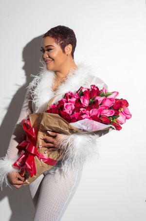 Woman with short purple hair holding large pink and red rose bouquet wrapped in kraft paper with red ribbon, wearing white feather coat.