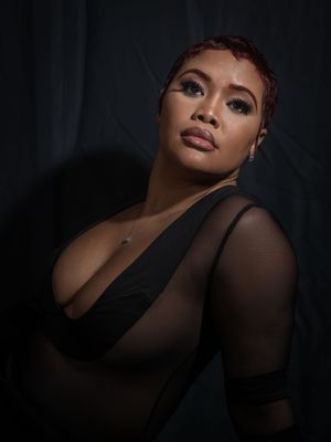 A woman with short red hair poses confidently in a black outfit against a dark backdrop in a dramatic studio portrait.