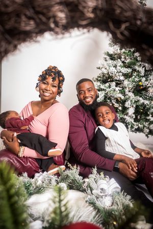 A joyful Black family poses together by a decorated Christmas tree, parents smiling with two young children in holiday attire.