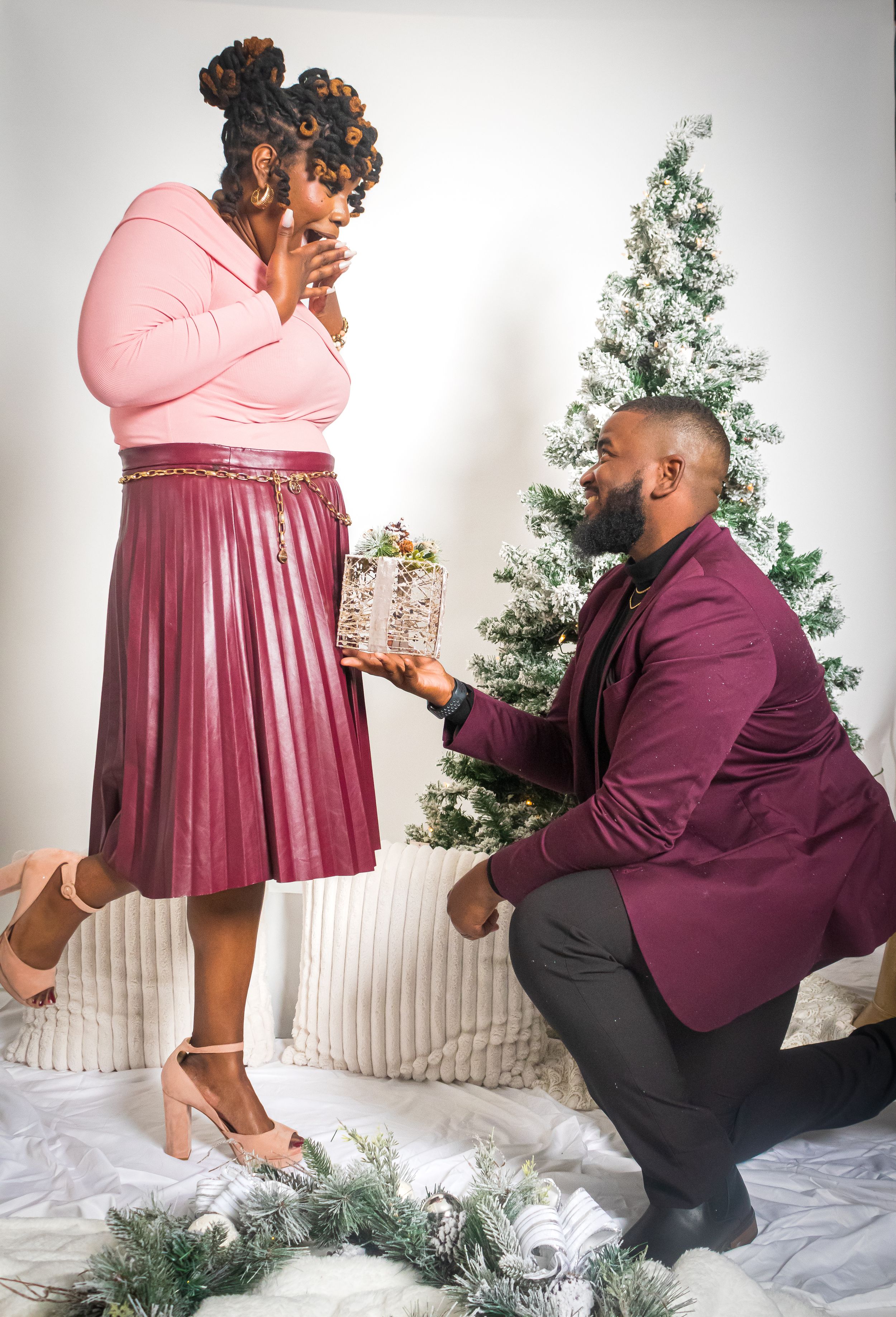 Man kneeling to present a gift to a surprised woman near a Christmas tree in a festive holiday photo.