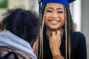 Smiling graduate in blue cap with colorful braids being congratulated by a loved one at graduation ceremony.