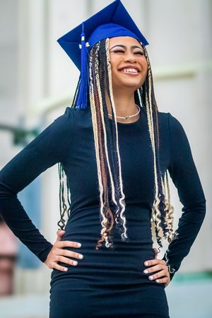 Smiling graduate in blue cap with long braids wearing black dress, posing confidently with hands on hips.