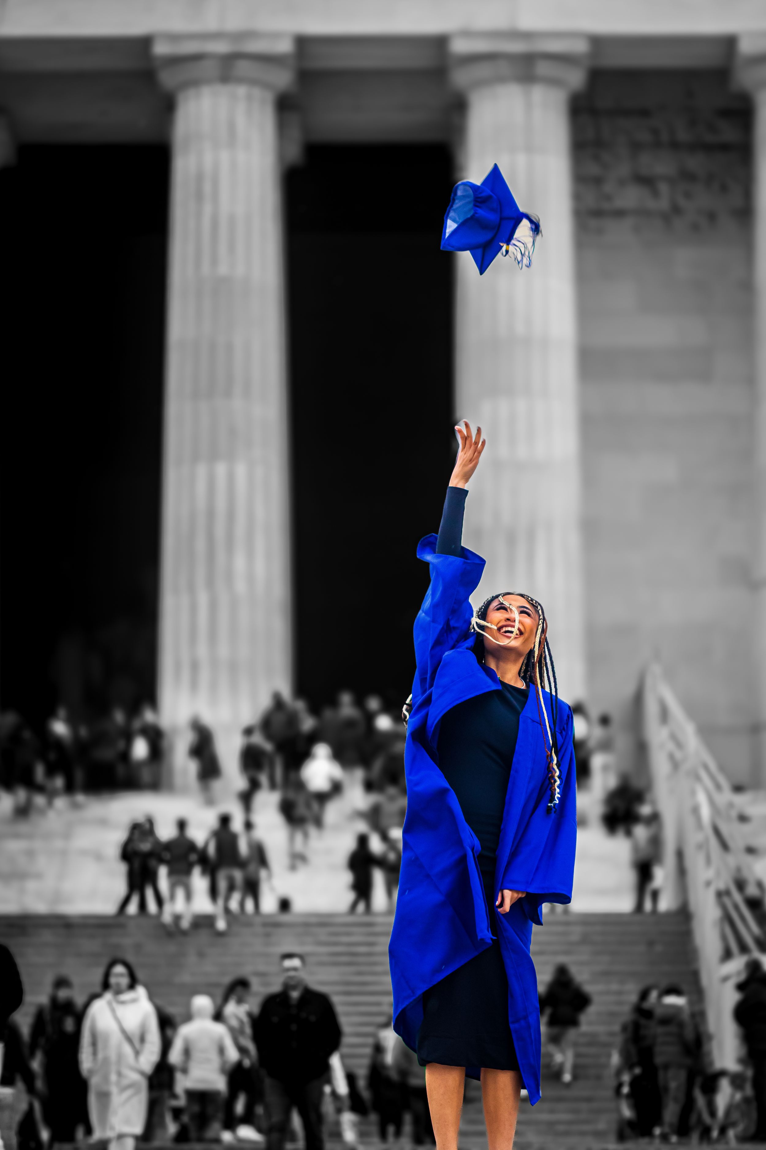Graduate in blue cap and gown tossing graduation cap in front of classical columns.