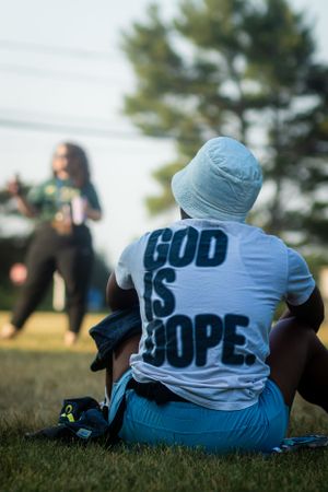 Person in light blue t-shirt with 'GOD IS DOPE' text sitting on grass during outdoor gathering.