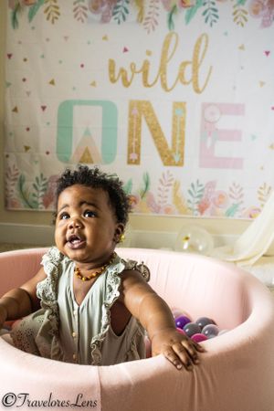 Smiling baby sitting in pink chair against floral 'Wild One' birthday backdrop.