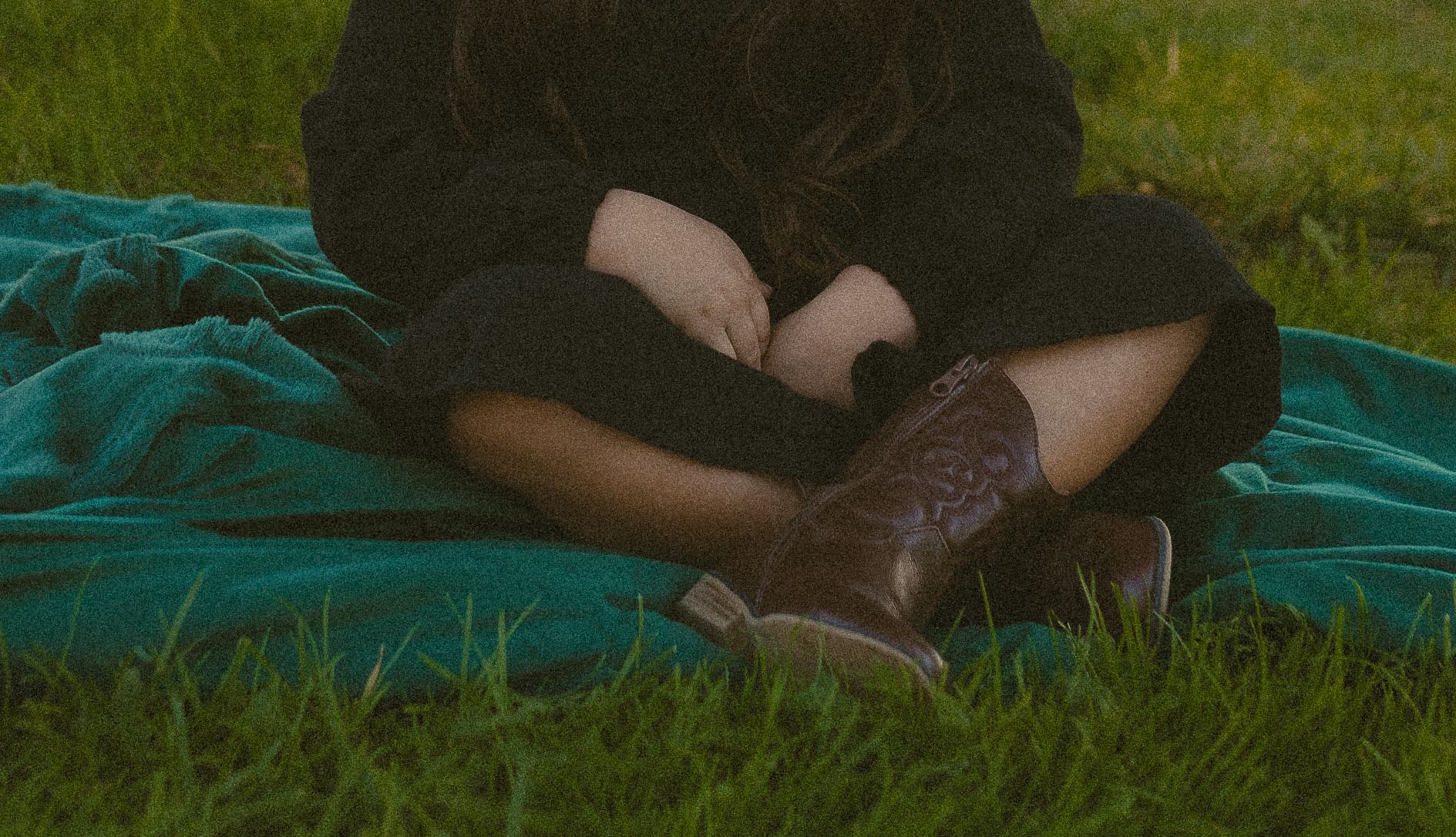 Person sitting on a teal blanket in the grass wearing boots and dark clothing.