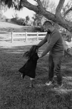 Parent and child playing and spinning together on grass near a wooden fence.