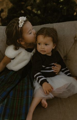 Two children sharing an affectionate moment on a couch, wearing winter clothing and a floral headband.