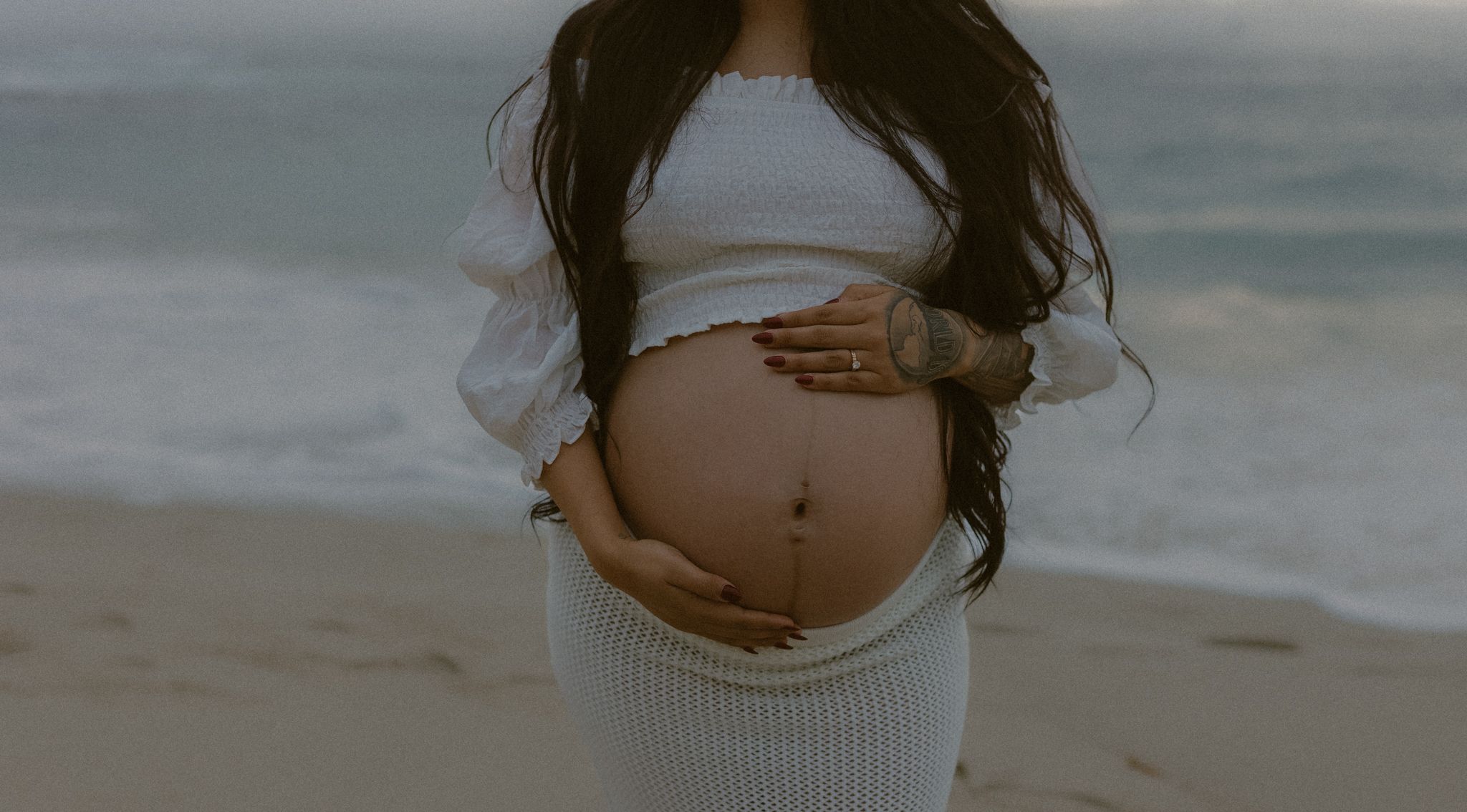 Expecting mother in a white sweater dress reveals her baby bump against an ocean backdrop.