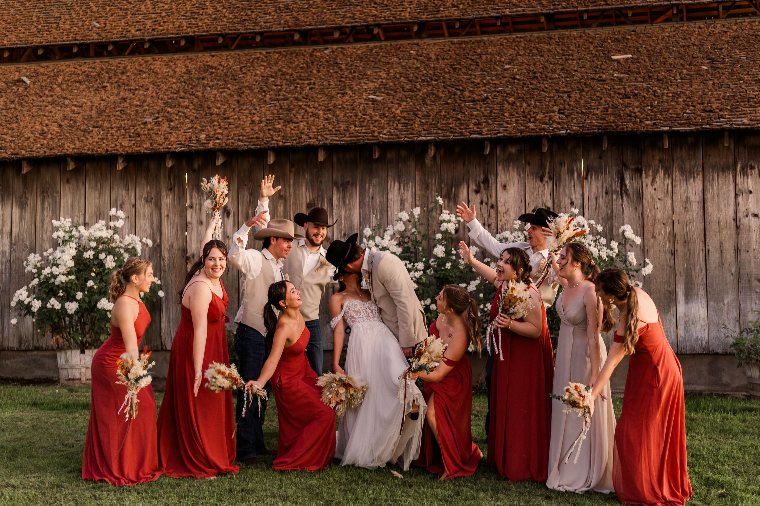 Wedding party in red bridesmaid dresses and white gown pose together in front of rustic barn setting.