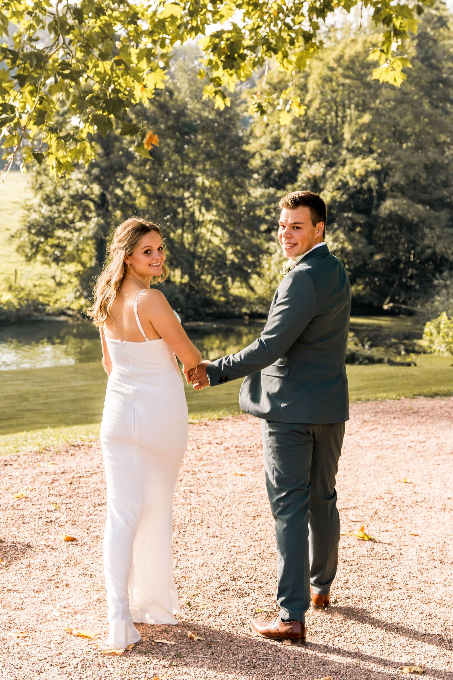 Sunlit autumn wedding scene with couple on gravel path near water.
