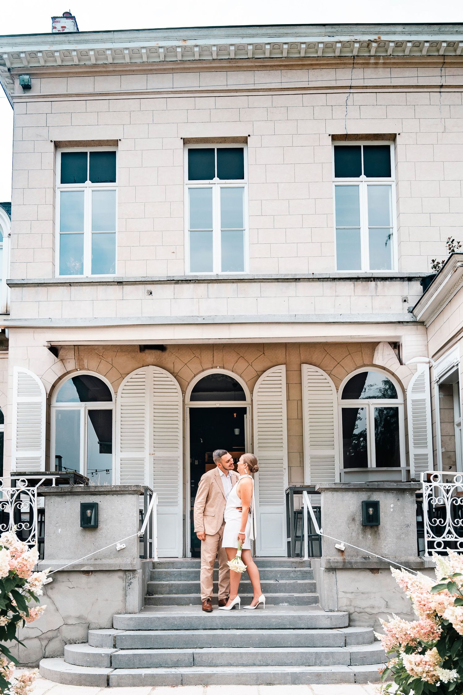 A couple stands on the steps of an elegant white villa with floral decorations.