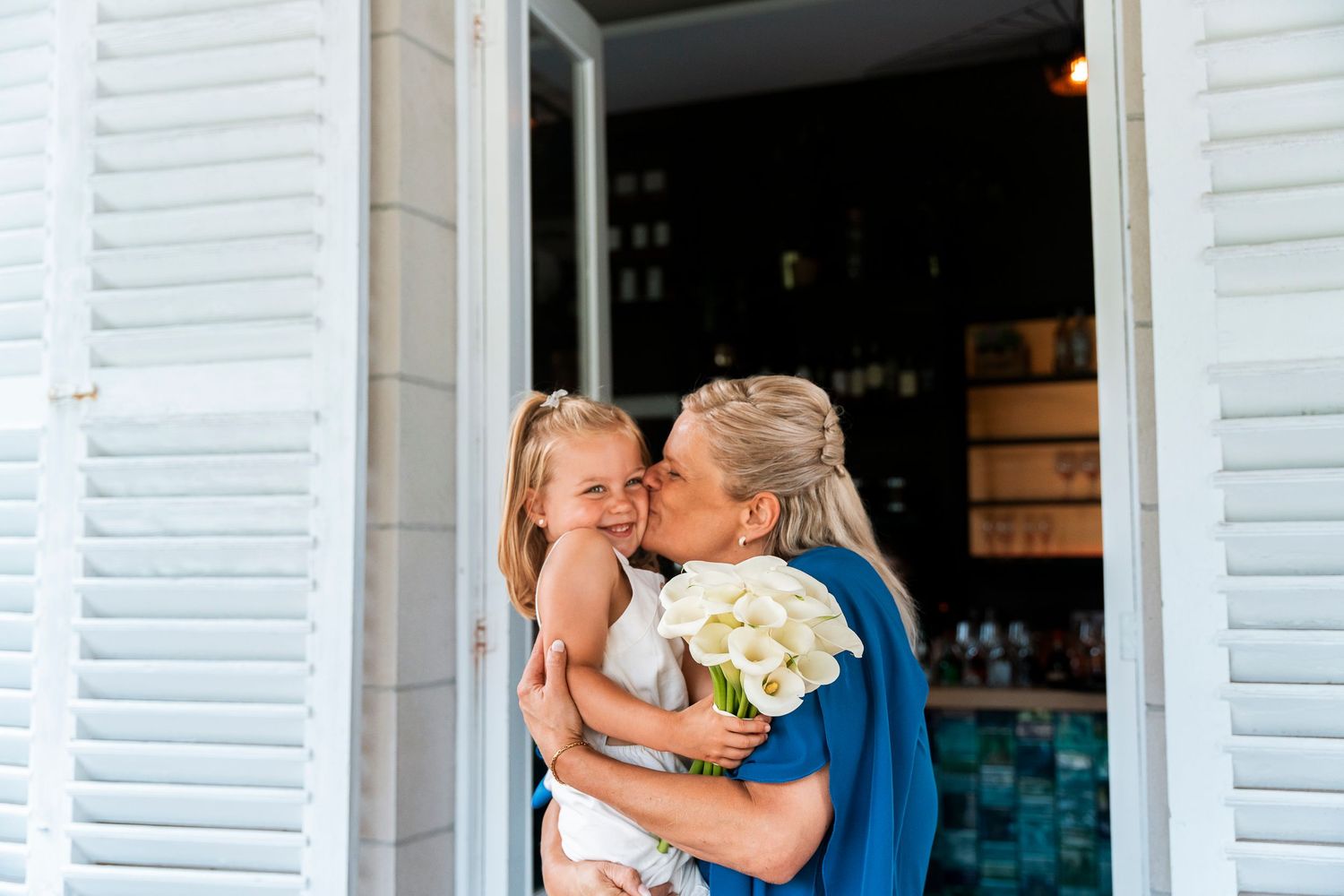Two people sharing a warm embrace while holding white flowers near white window shutters.
