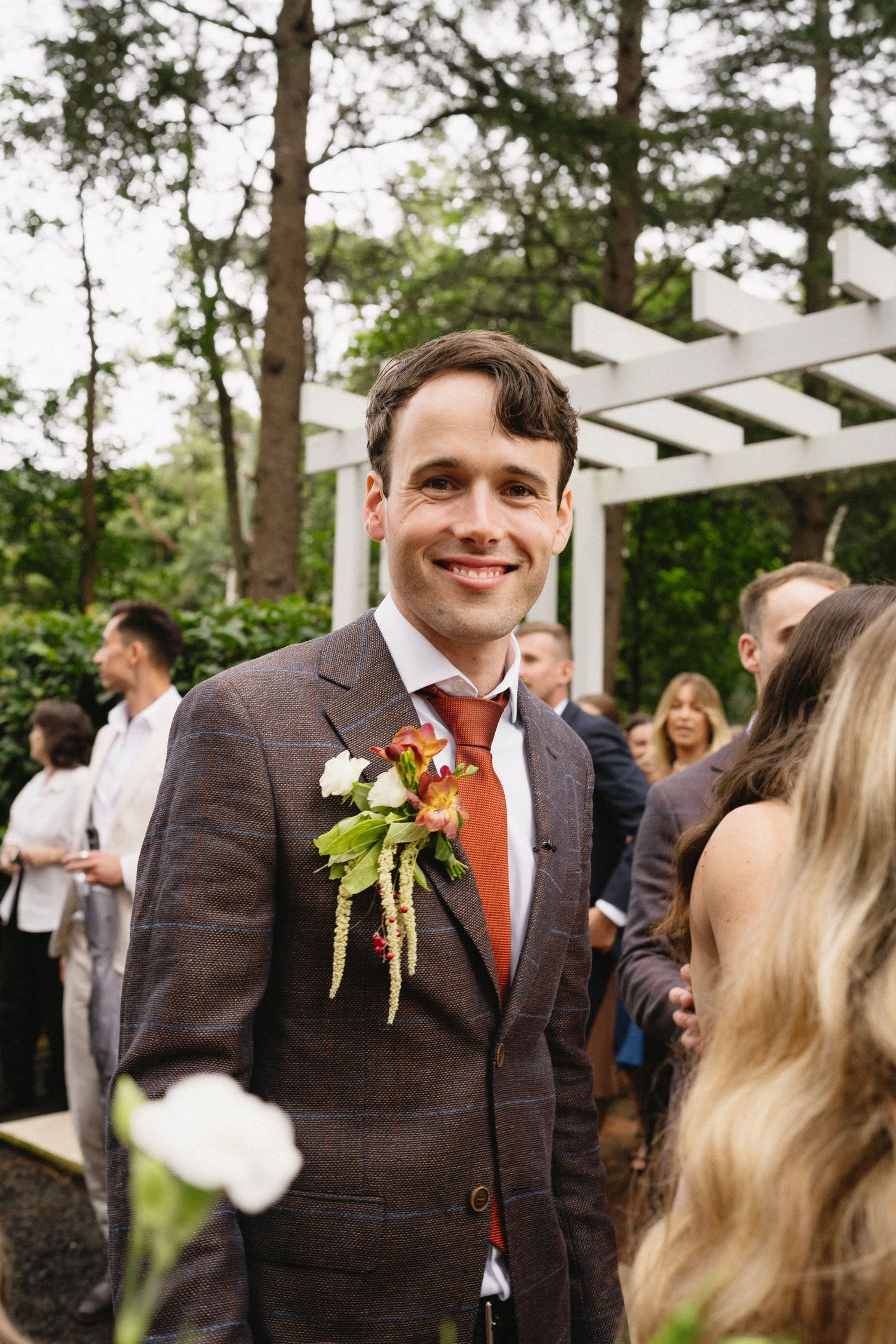 Groom in brown suit and red tie smiles during outdoor wedding reception.