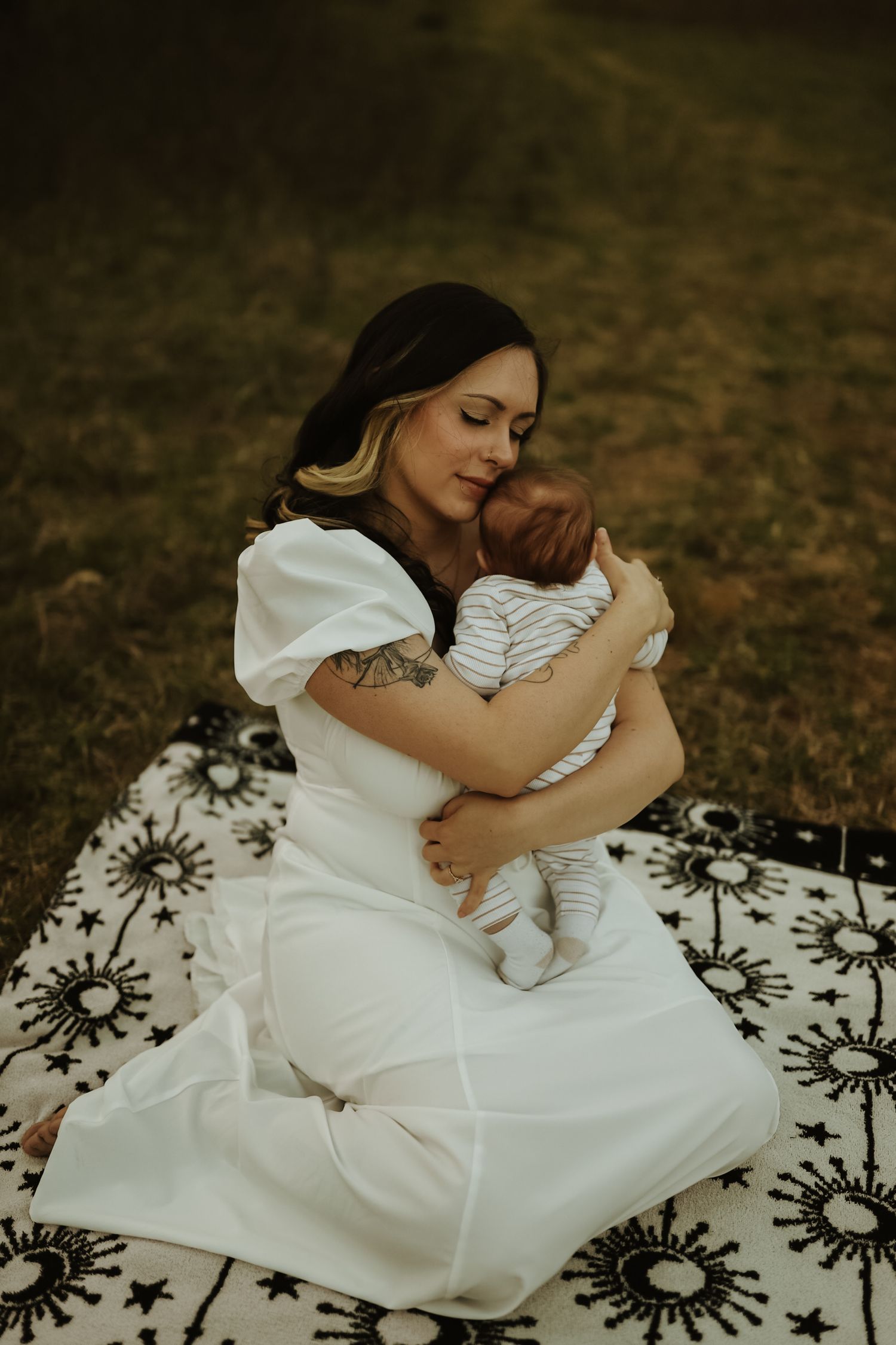 A mother in a white dress tenderly holding a newborn baby on a patterned blanket outdoors.