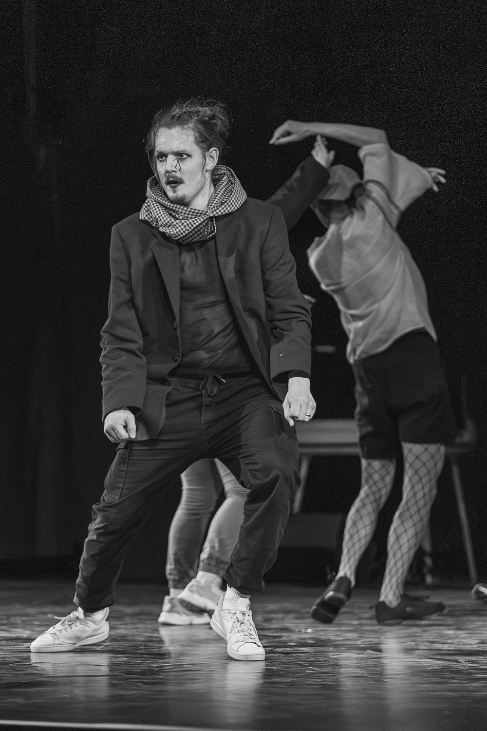 Black and white photo of performers dancing expressively on stage, one in a scarf and coat, another in motion behind.