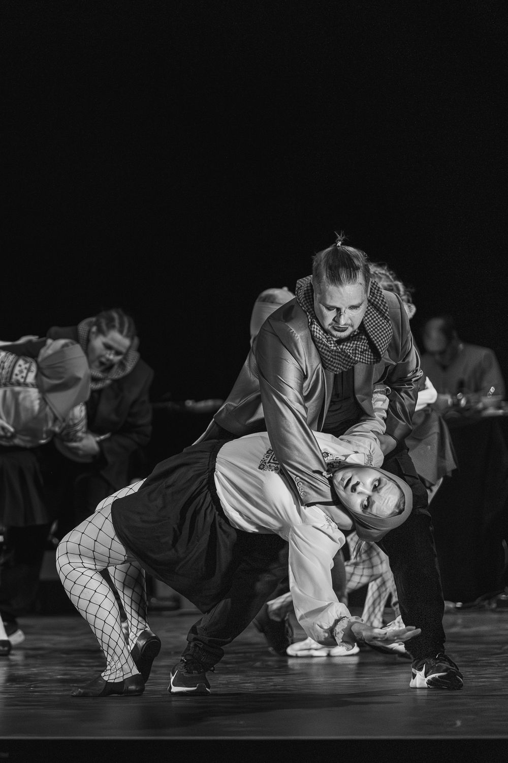 Two dancers perform an intense contemporary dance move in dramatic black and white lighting on a dark stage.