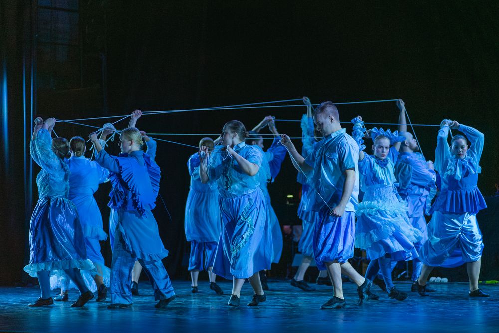 Performers in long flowing dresses dance together on stage bathed in dramatic blue lighting during a theatrical production.