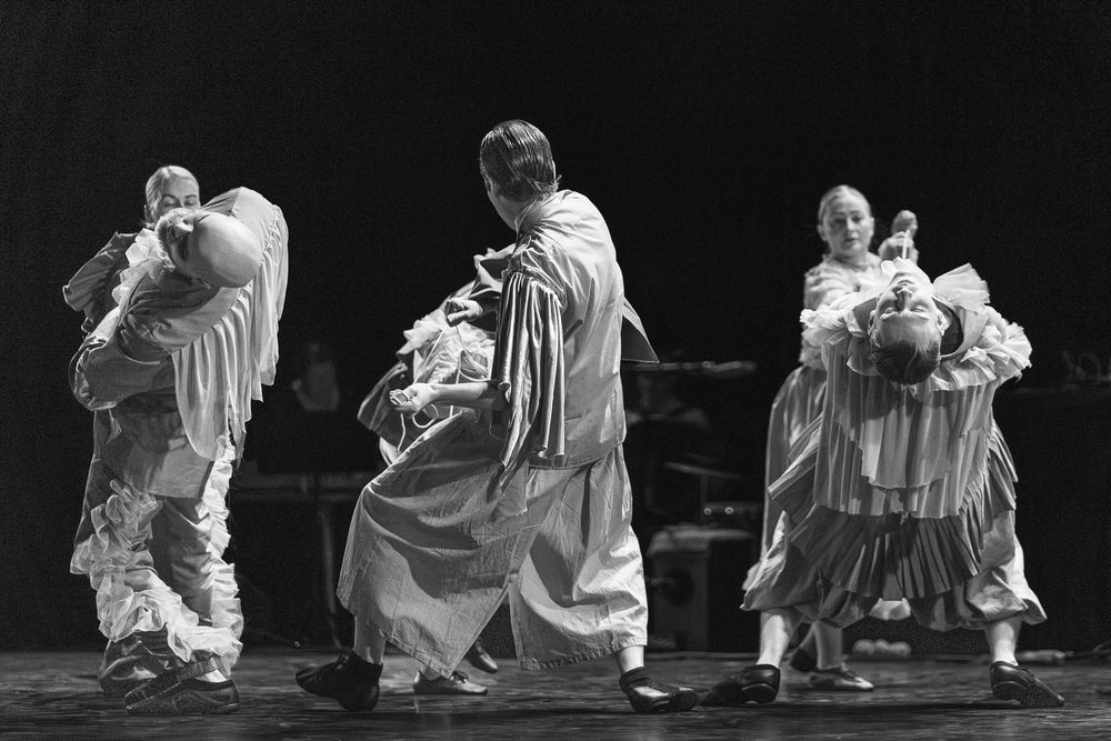 Black and white photograph of performers in flowing white robes dancing together on a dark stage in an expressive movement.