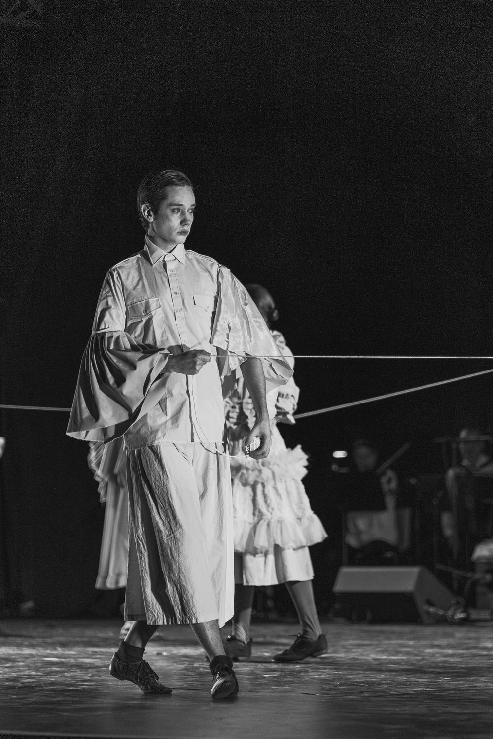 Black and white stage performance photograph shows a performer in flowing white robes moving across a darkened stage.