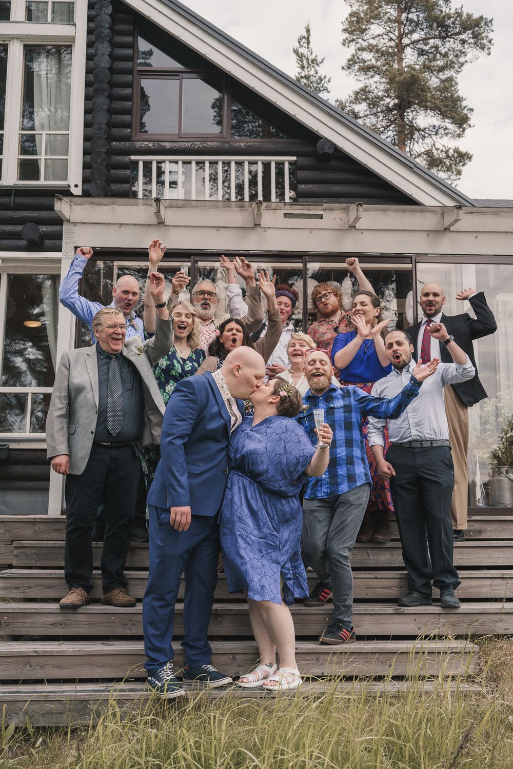 A cheerful group celebrates together on the porch steps of a rustic wooden house with an A-frame design.