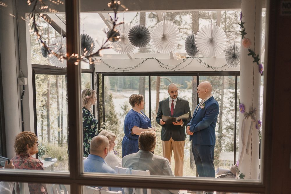Beautifully decorated indoor venue with glass walls showing trees outside while people gather for an elegant event.