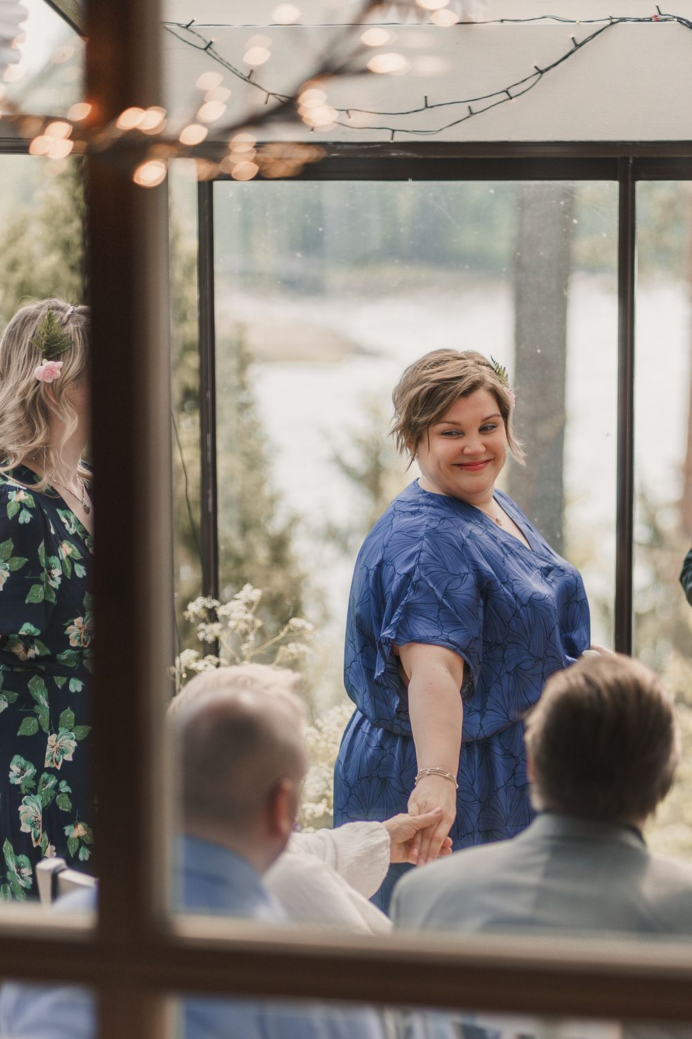 A person wearing a blue dress smiles joyfully during an outdoor gathering seen through a window.
