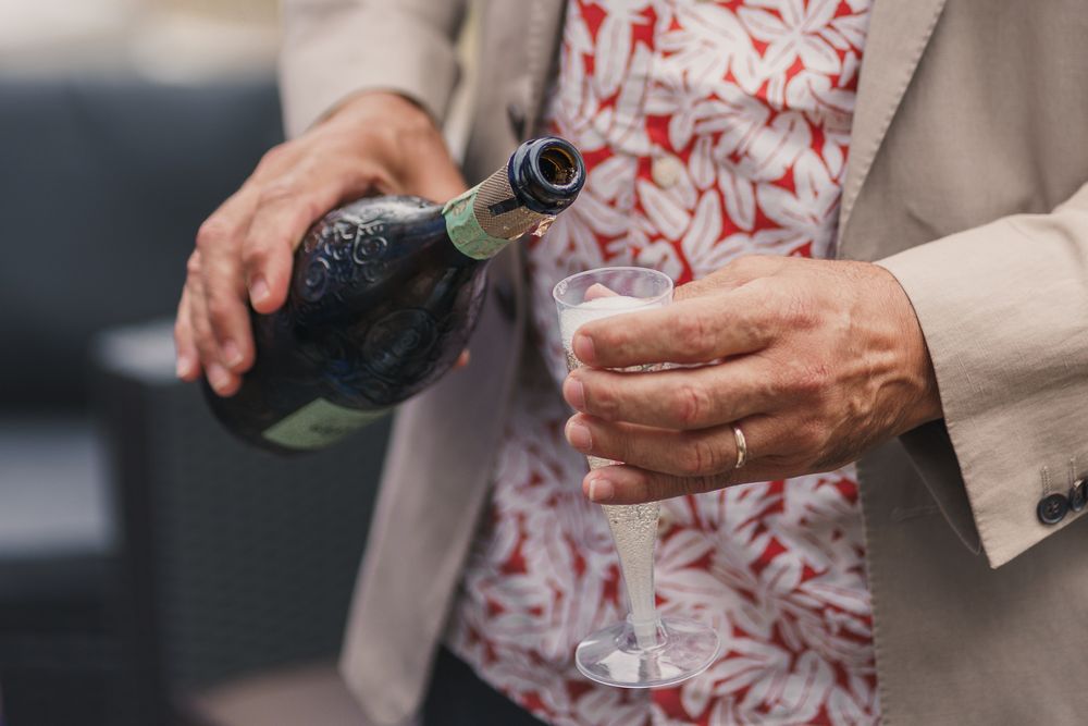 Hands pouring champagne into a glass during a celebration, wearing beige jacket with red and white floral pattern.