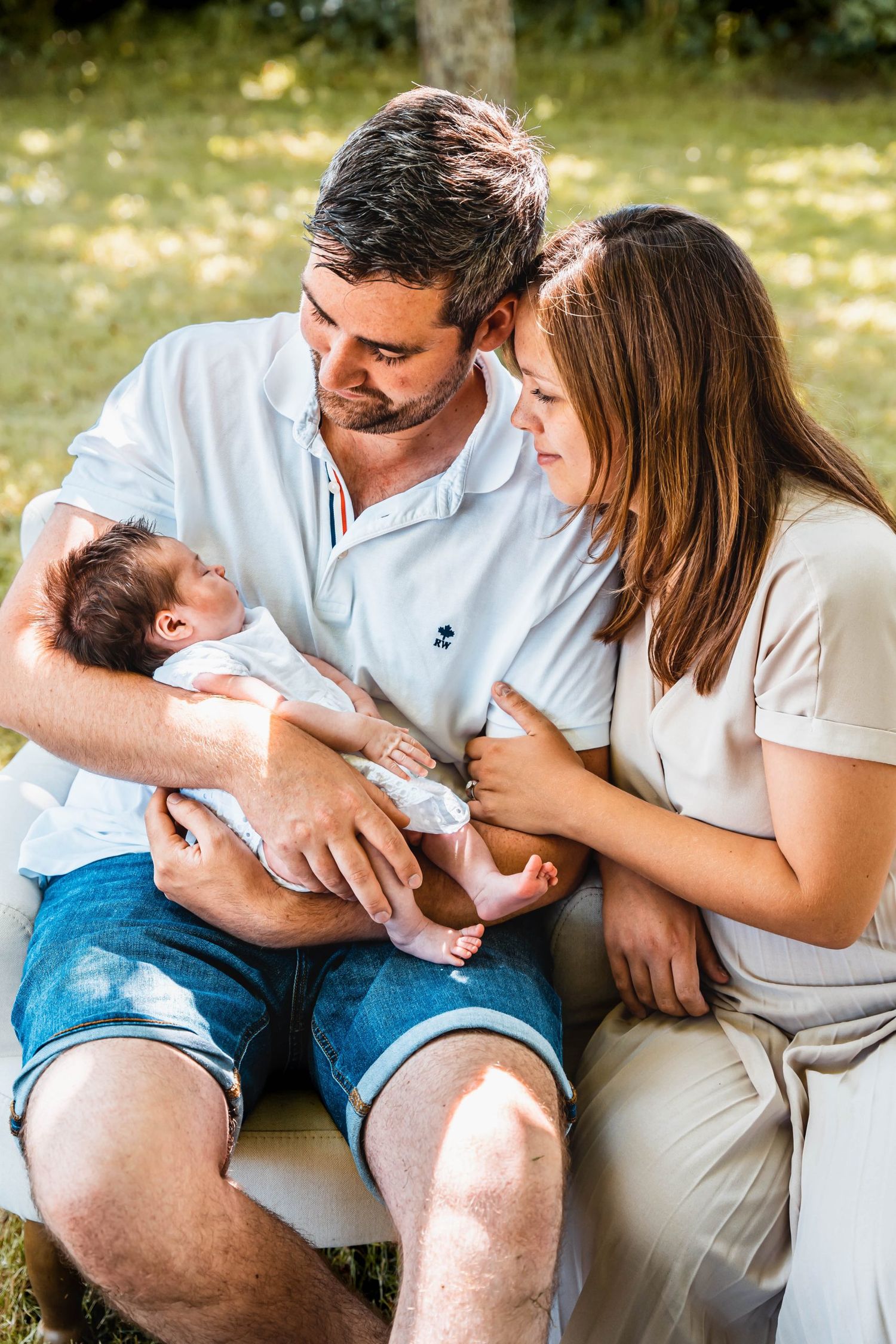 Family sharing intimate moment with newborn baby outdoors on white chair.