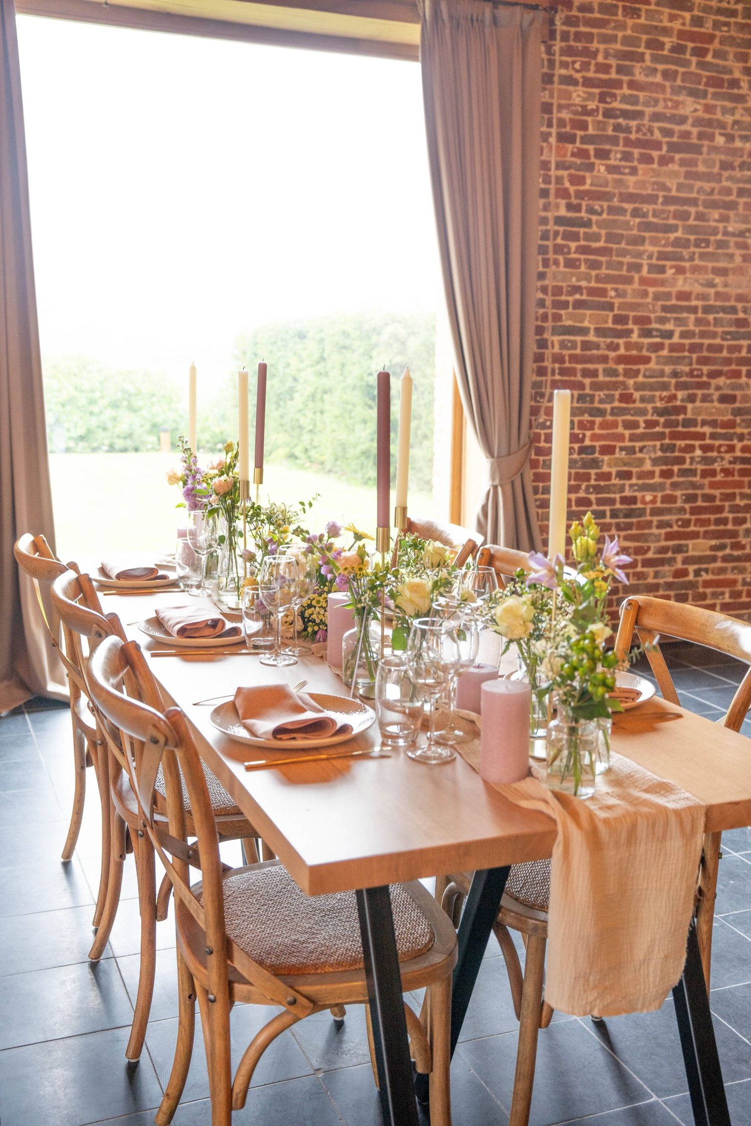 Elegant table setting with wooden chairs, exposed brick wall, and fresh floral centerpieces in glass vases.