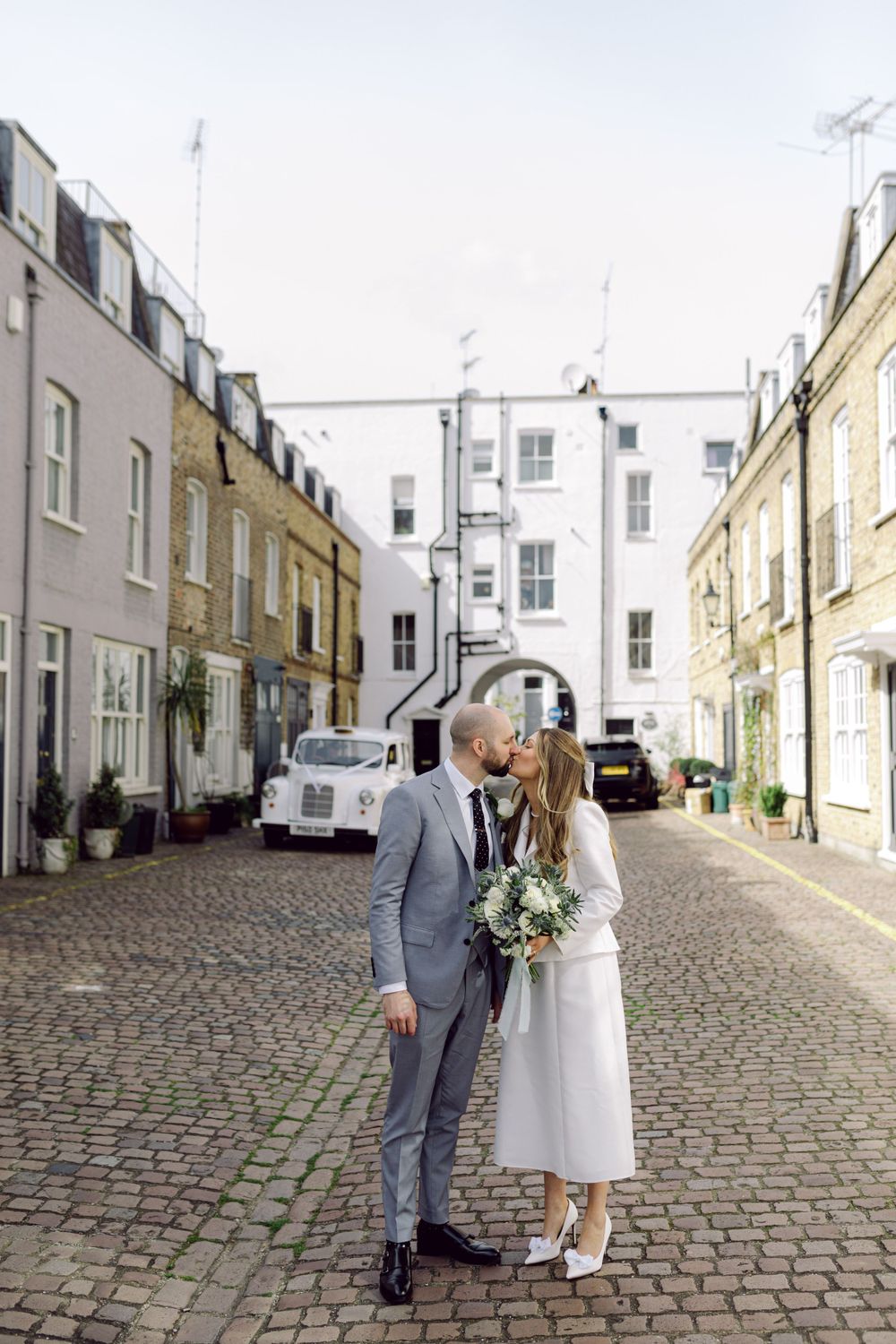 A couple in wedding attire share a moment on a charming cobblestone street lined with pastel townhouses in London.