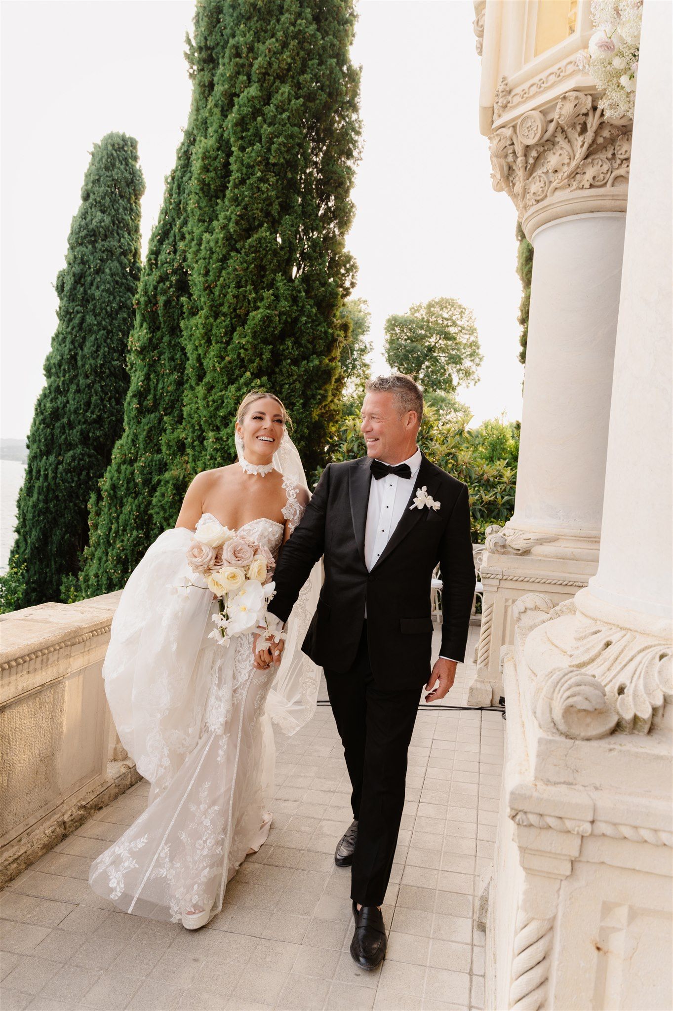 Newlyweds walk together along elegant villa terrace with tall cypress trees in background.