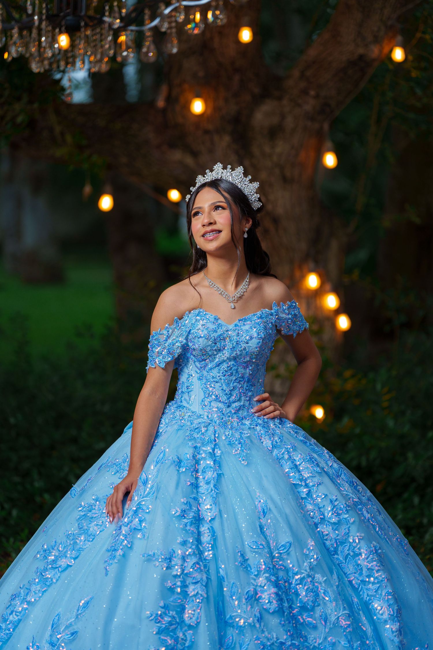 An elegant blue ball gown with off-shoulder design photographed outdoors with twinkling lights in the background.