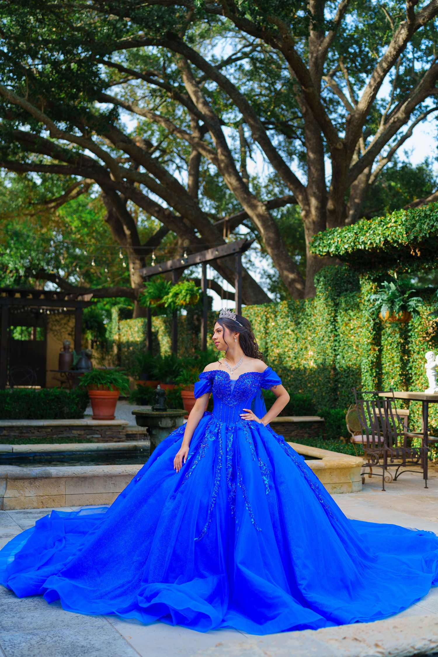 A royal blue ball gown with dramatic train spreads across a garden patio beneath large oak trees.