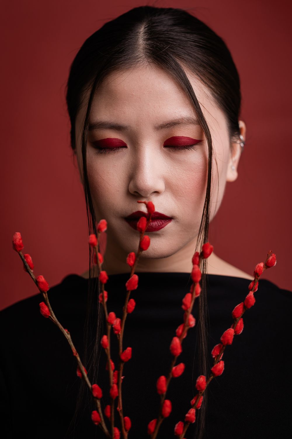 Artistic fashion portrait with red flower branches against dark red background featuring dramatic eye makeup.