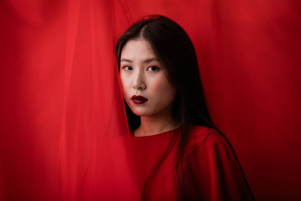 A dramatic portrait against a red background features striking dark lipstick and long black hair in a monochromatic style.