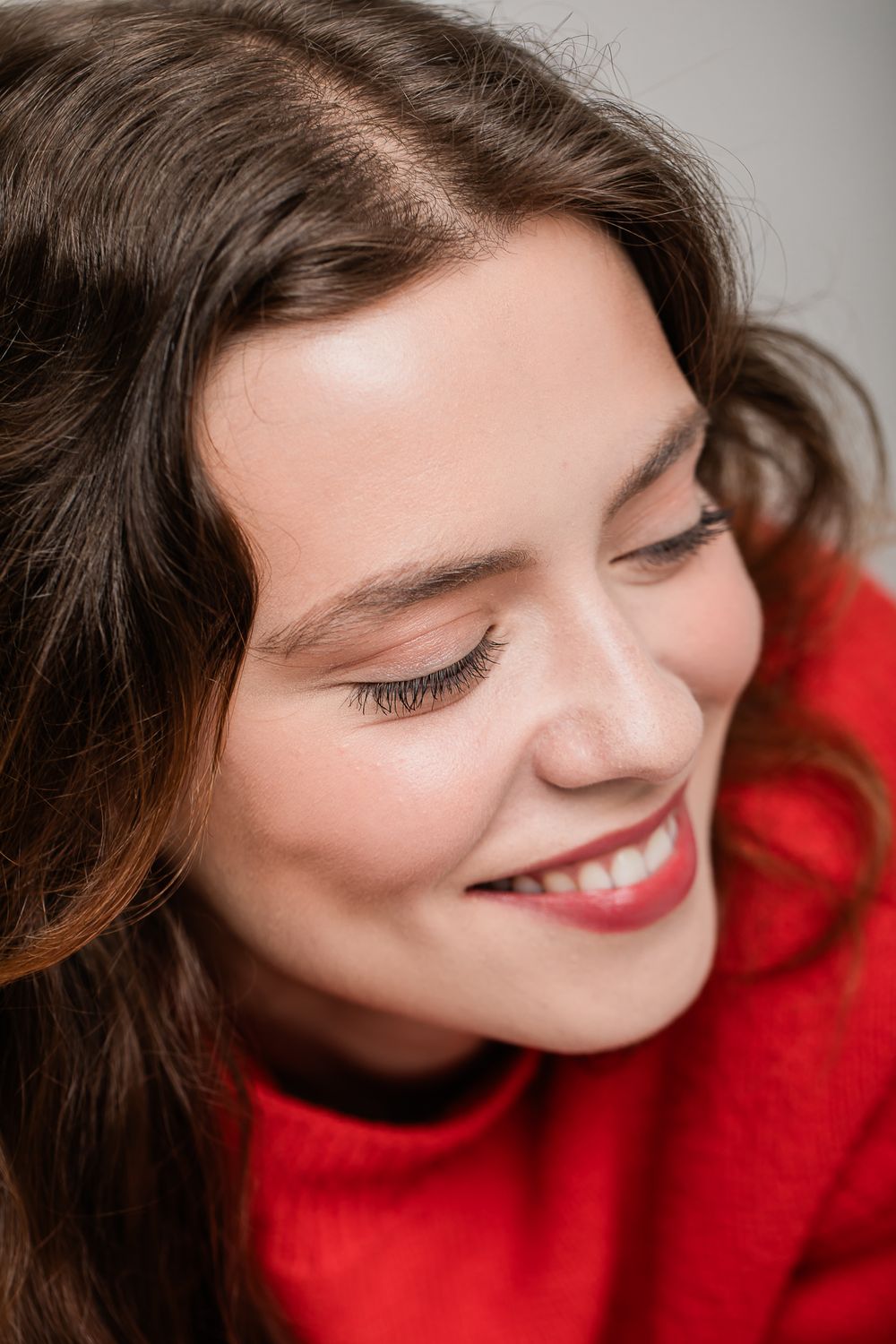 Portrait photo showing a joyful smile, closed eyes, brown wavy hair, and vibrant red sweater in a candid moment.