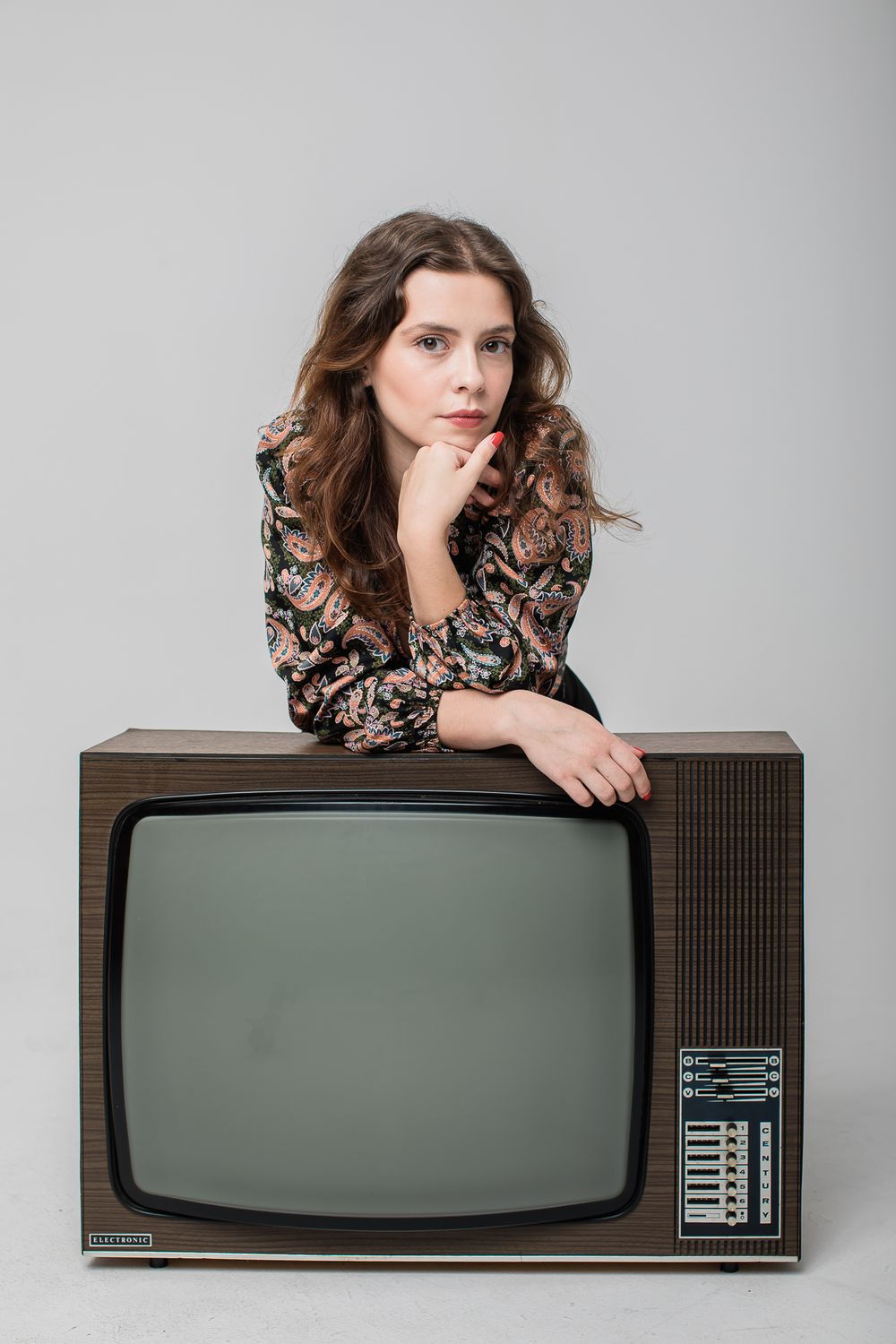 Person in floral blouse leaning on vintage television set against gray background in studio portrait.