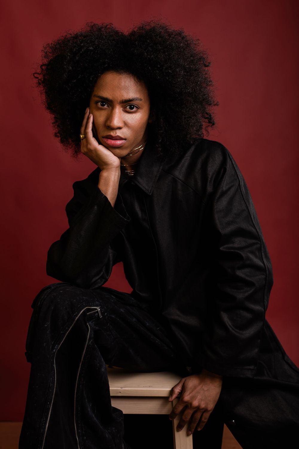 Studio portrait with natural curly hair in black outfit against dark red background posing thoughtfully.