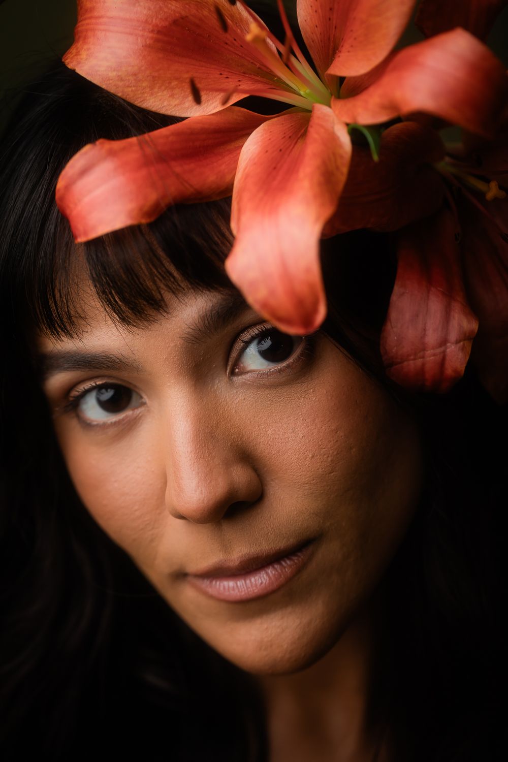 A striking portrait with dark hair and dramatic coral-colored lily flower against a black background.