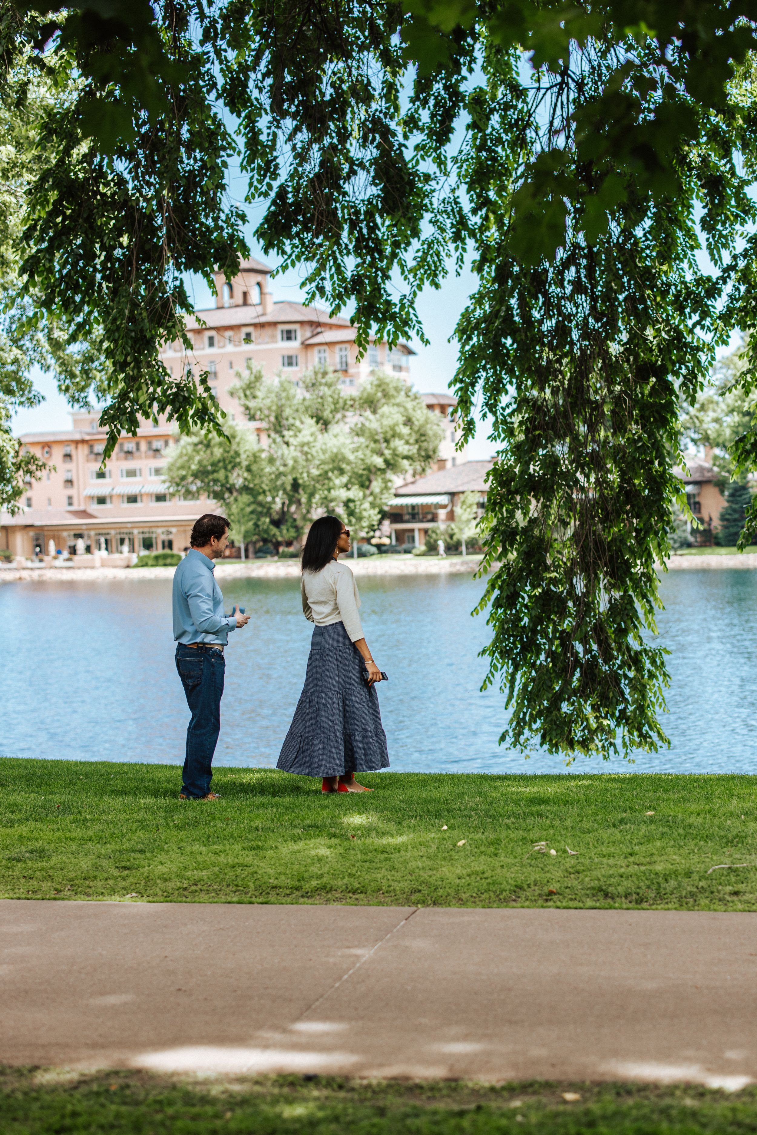 Two figures stand beneath a weeping willow tree by a serene lakeside on a sunny summer afternoon.