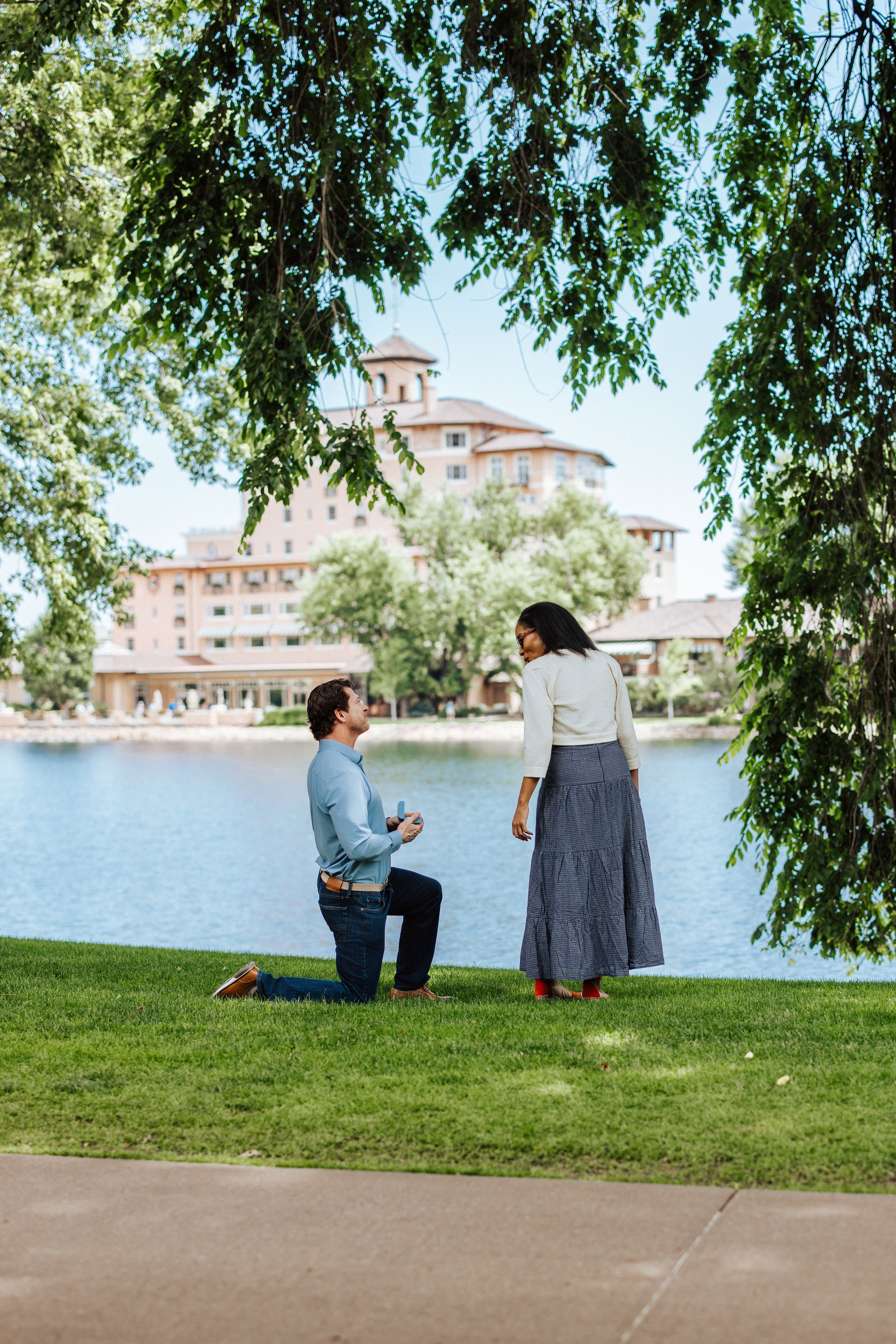 Two people share a quiet moment sitting on a grassy hill overlooking a scenic lake with buildings in the background.
