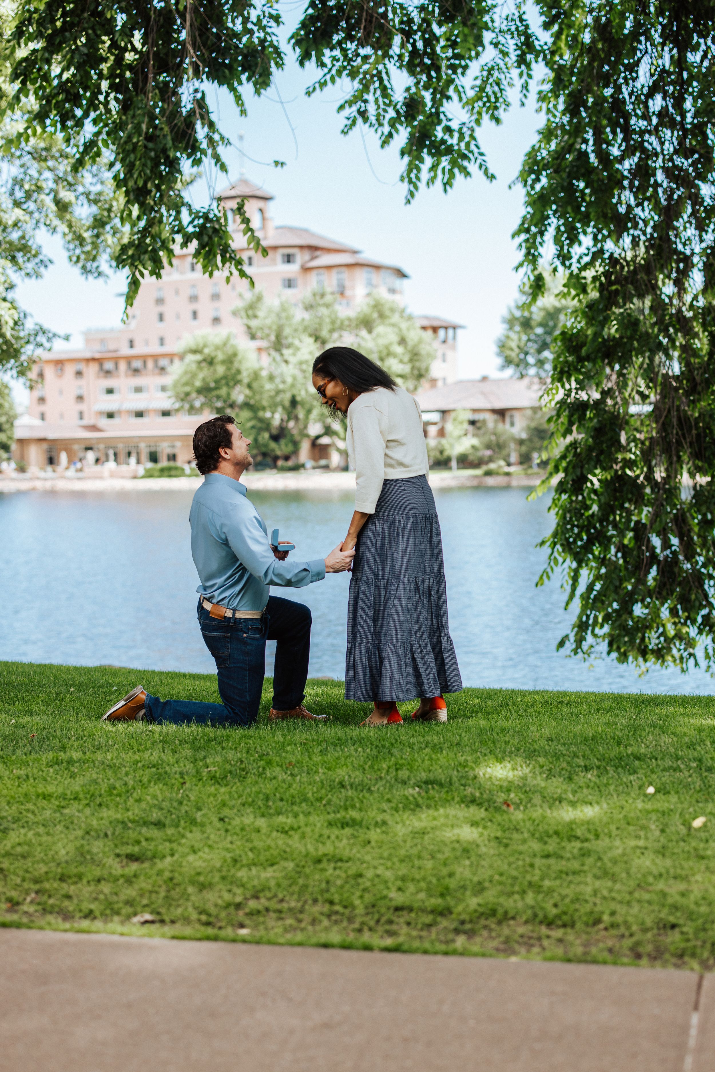A couple sits together on a grassy hill overlooking a peaceful lake on a sunny summer day.