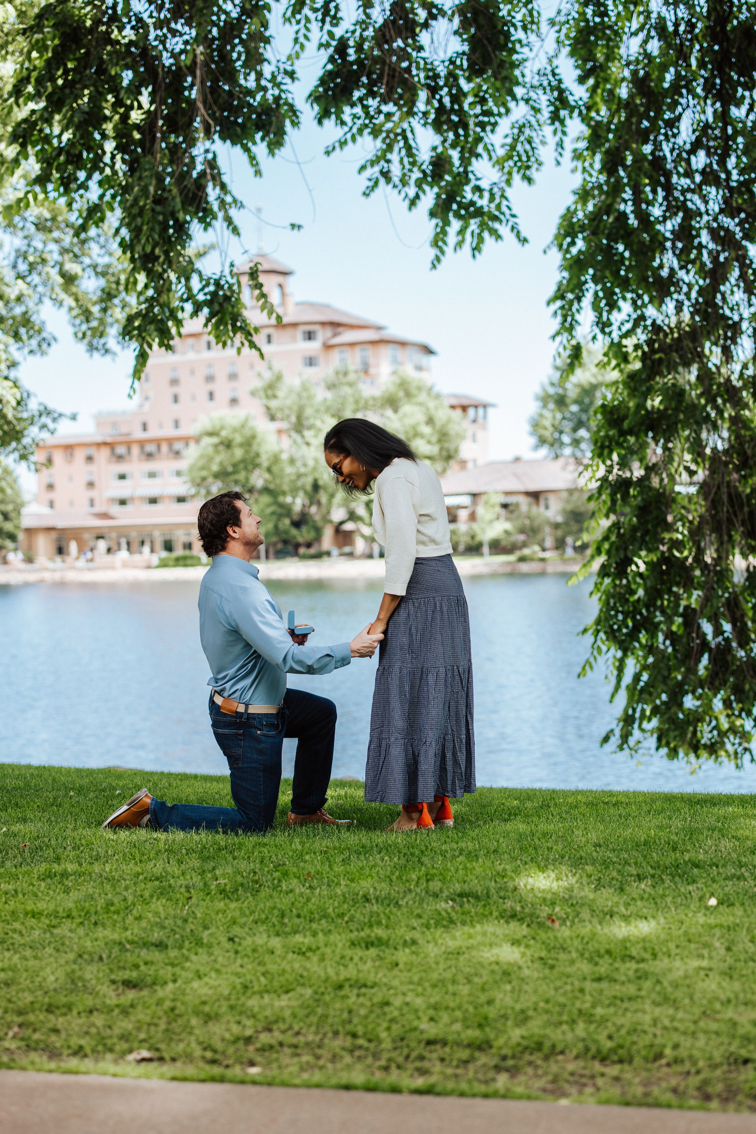 A special moment captured during a marriage proposal next to a lakeside building framed by tree branches.