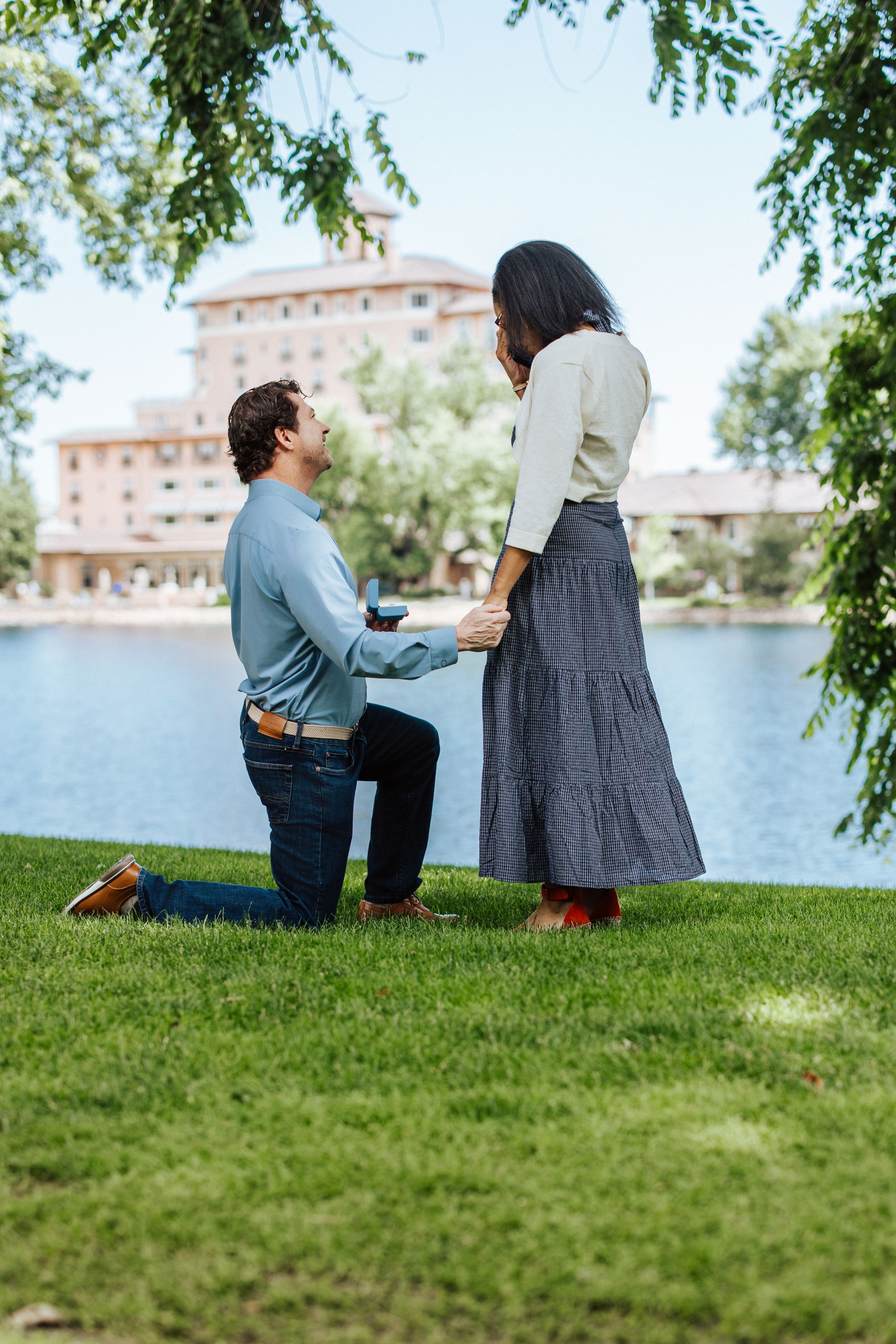A romantic proposal scene by a lake with historic architecture in the background on a sunny day.