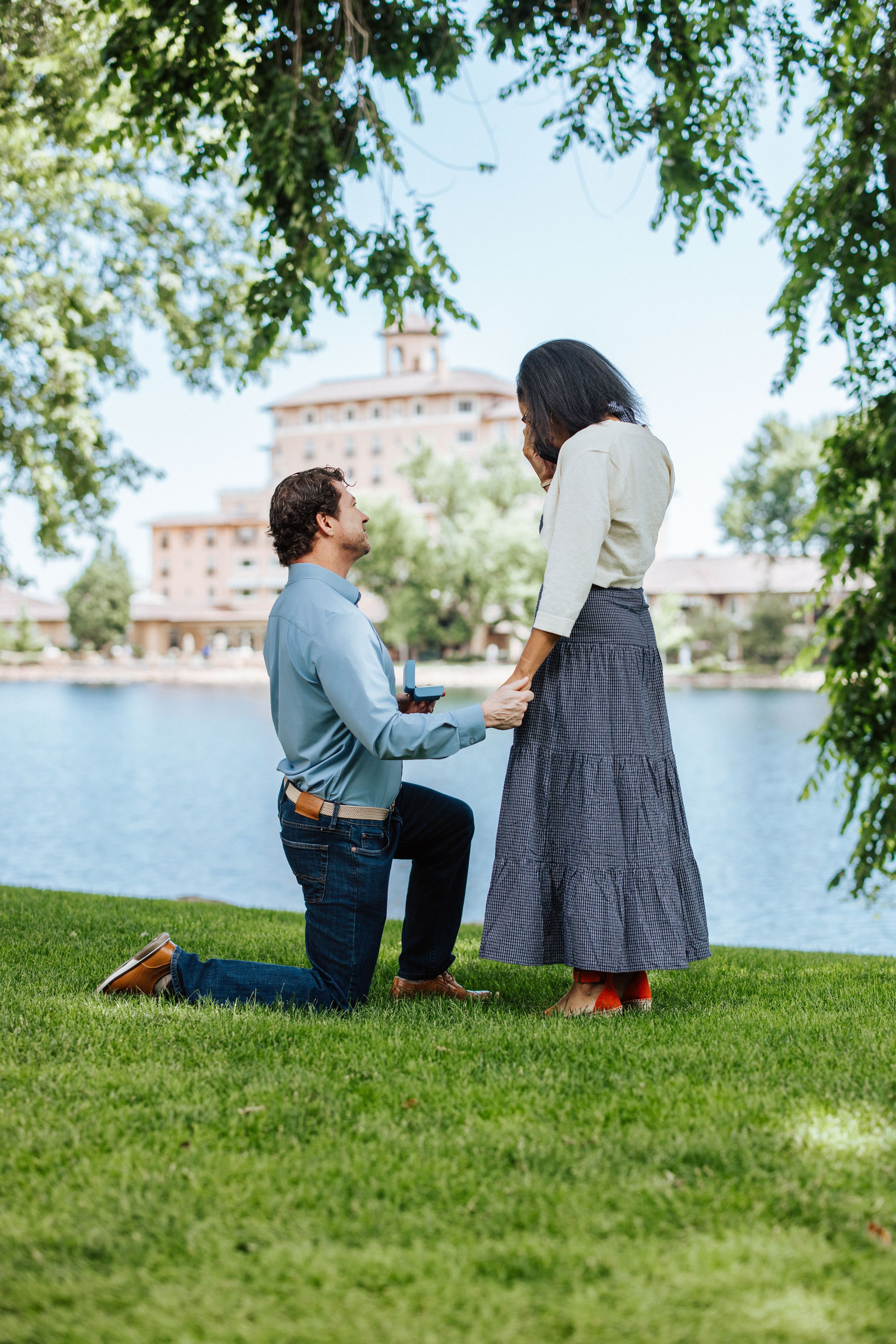 A romantic proposal scene unfolds on a grassy hill by a lake with buildings in the background.