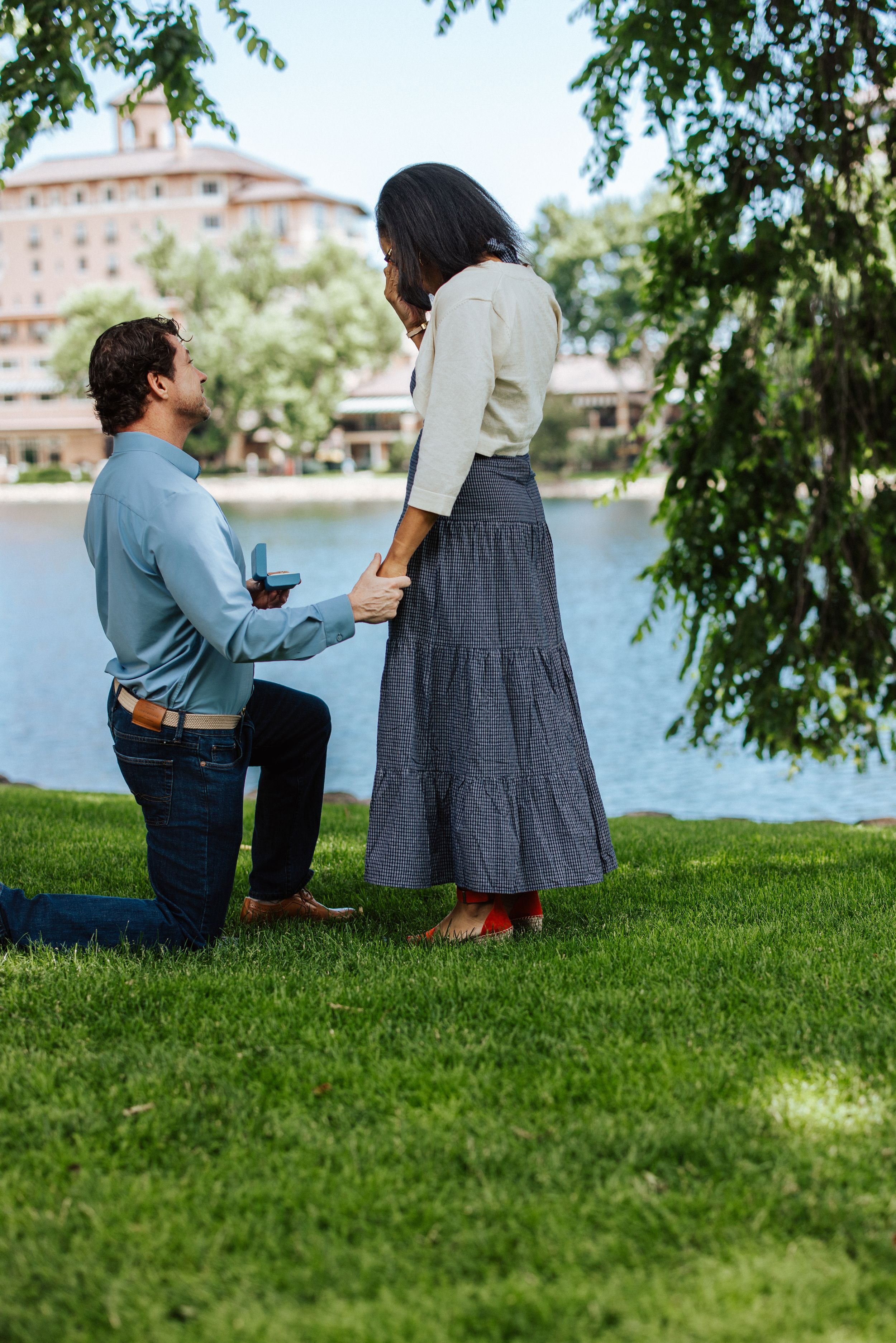 An intimate proposal moment on a grassy lawn beside a waterfront with a grand building in the distance.