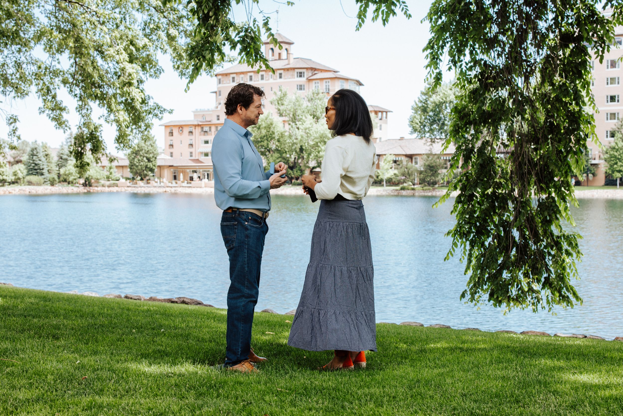 A couple stands together on a grassy lawn near a lake with a building visible in the background.