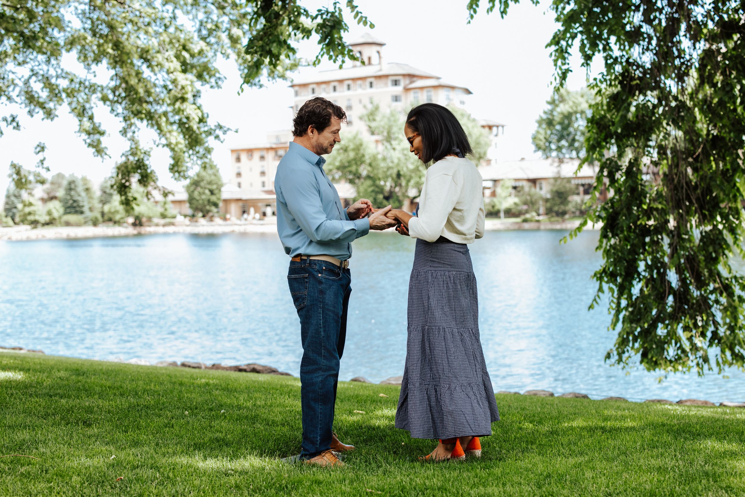 A couple stands beneath green tree branches near a serene waterfront setting in a romantic outdoor moment.