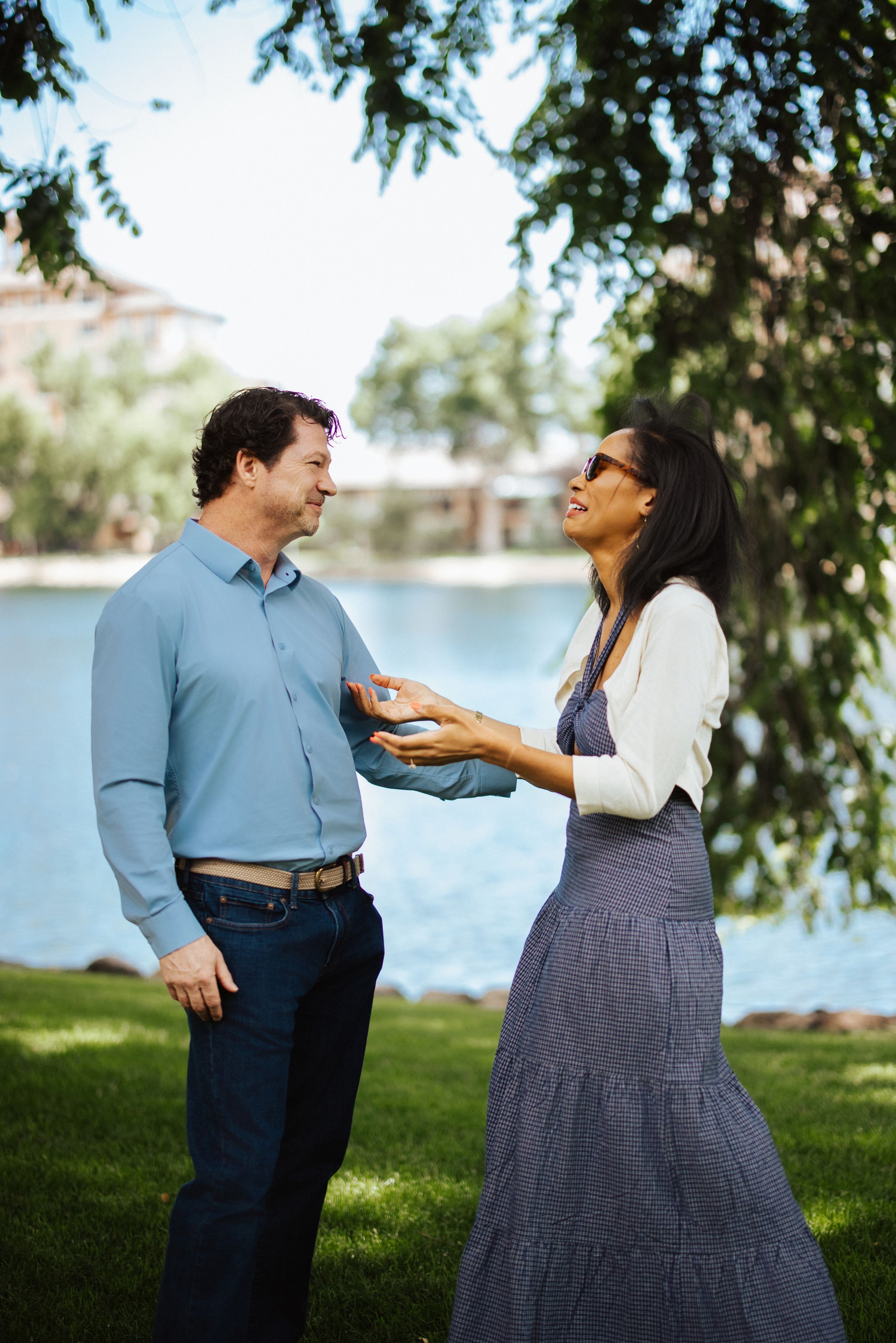 A couple shares a playful moment while laughing together on grass near a lake during a sunny day.